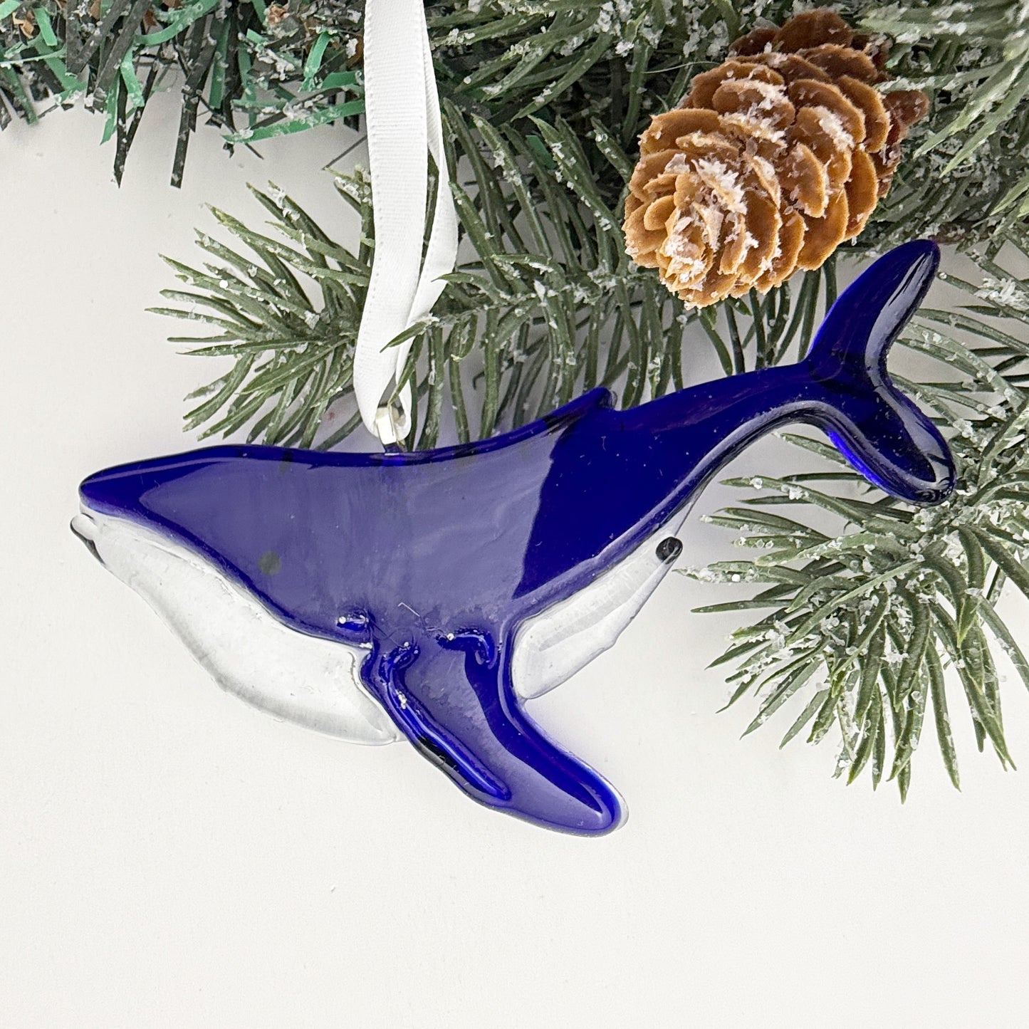 Transparent blue whale Christmas ornament in fused glass, with tree lights sparkling through, ocean-inspired holiday décor.
