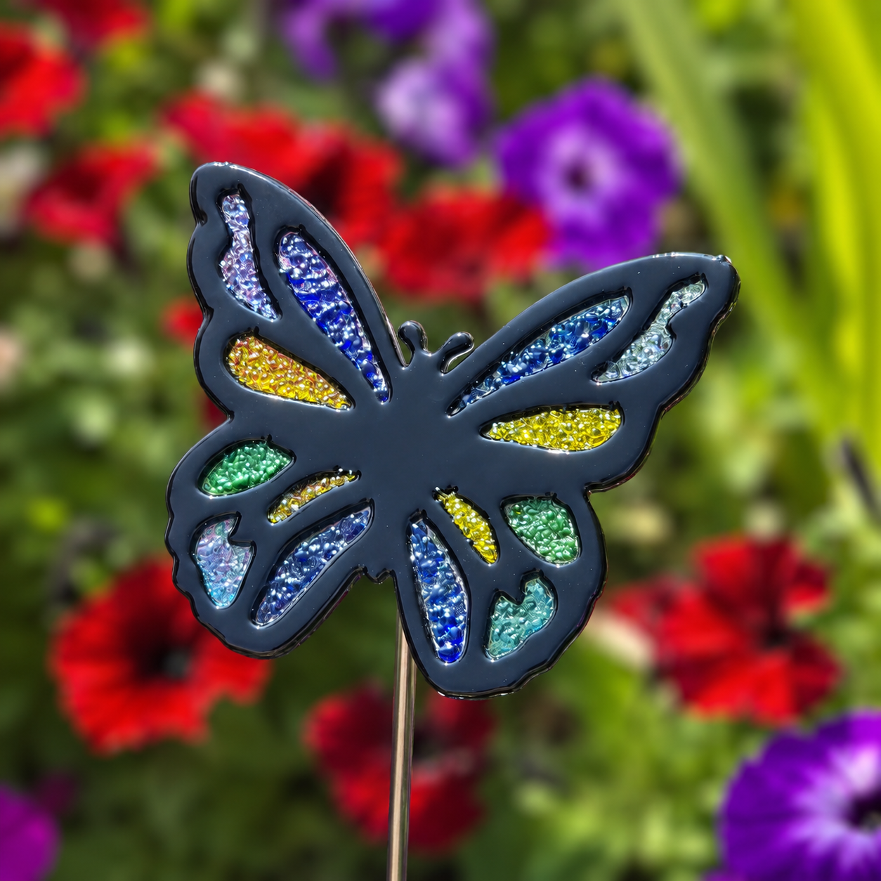 Fused glass butterfly garden stake with black outline and blue, green and yellow frit accents among flowers.