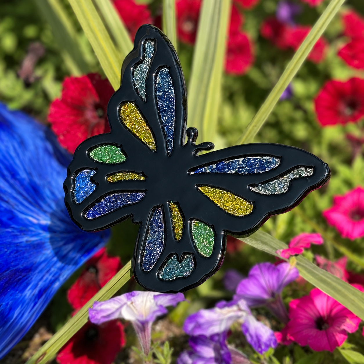 Finished fused glass butterfly garden stake with black outline and blue, green and yellow frit accents among flowers.
