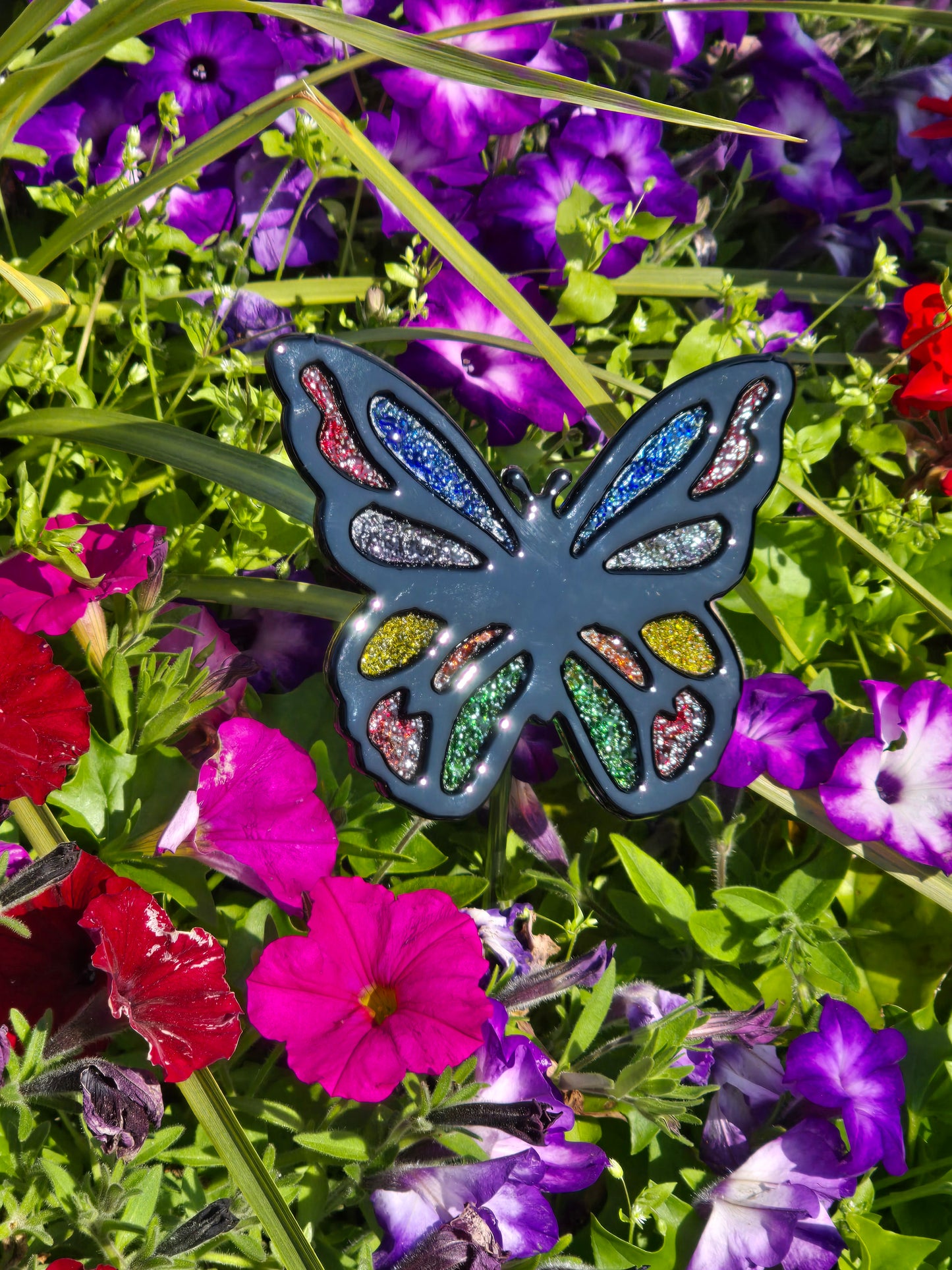Finished fused glass butterfly garden stake with multicolour frit accents displayed among purple and pink flowers.