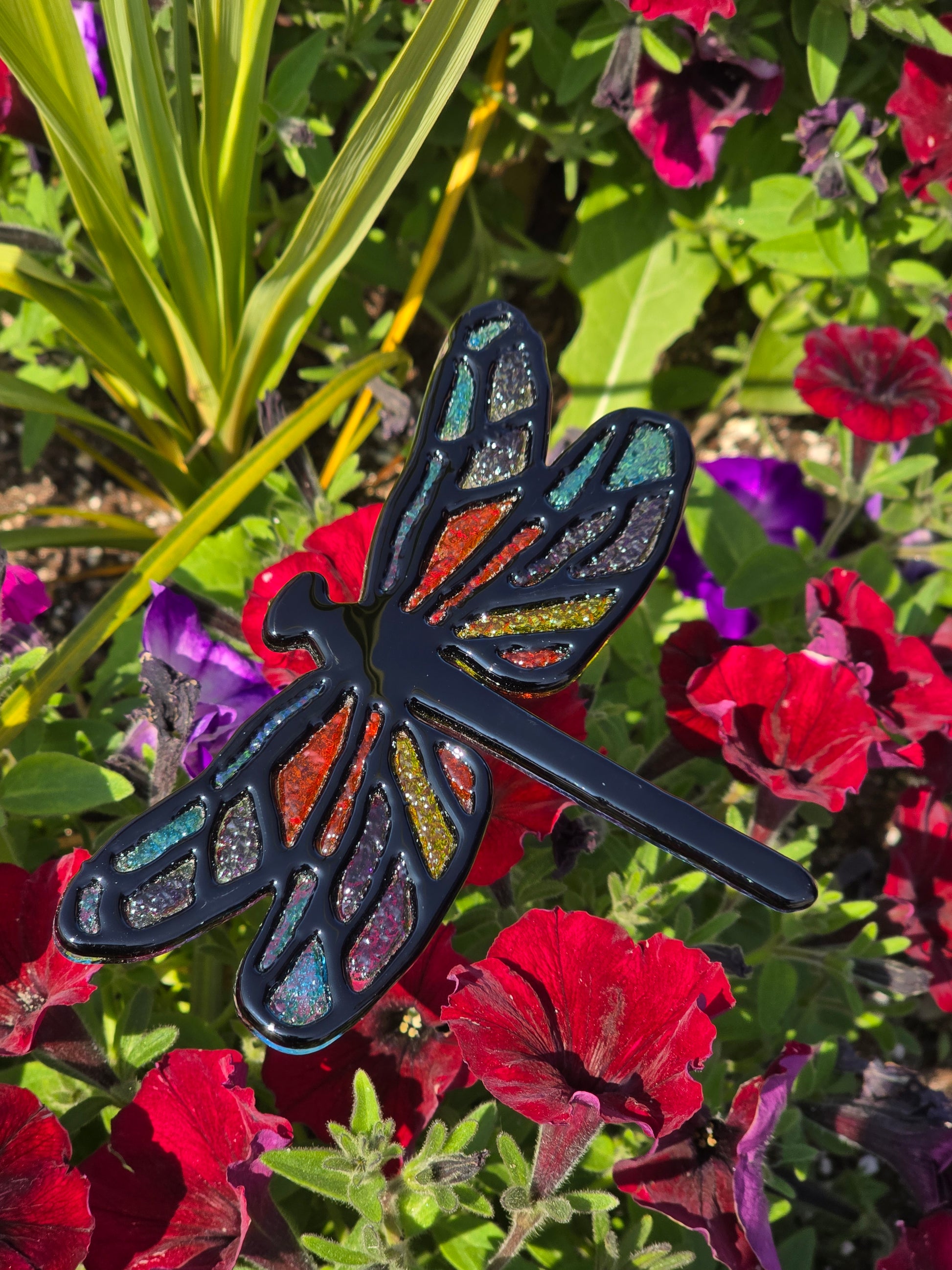A black fused glass dragonfly garden stake with multicolor frit wings, shown in a garden among red flowers and green leaves.