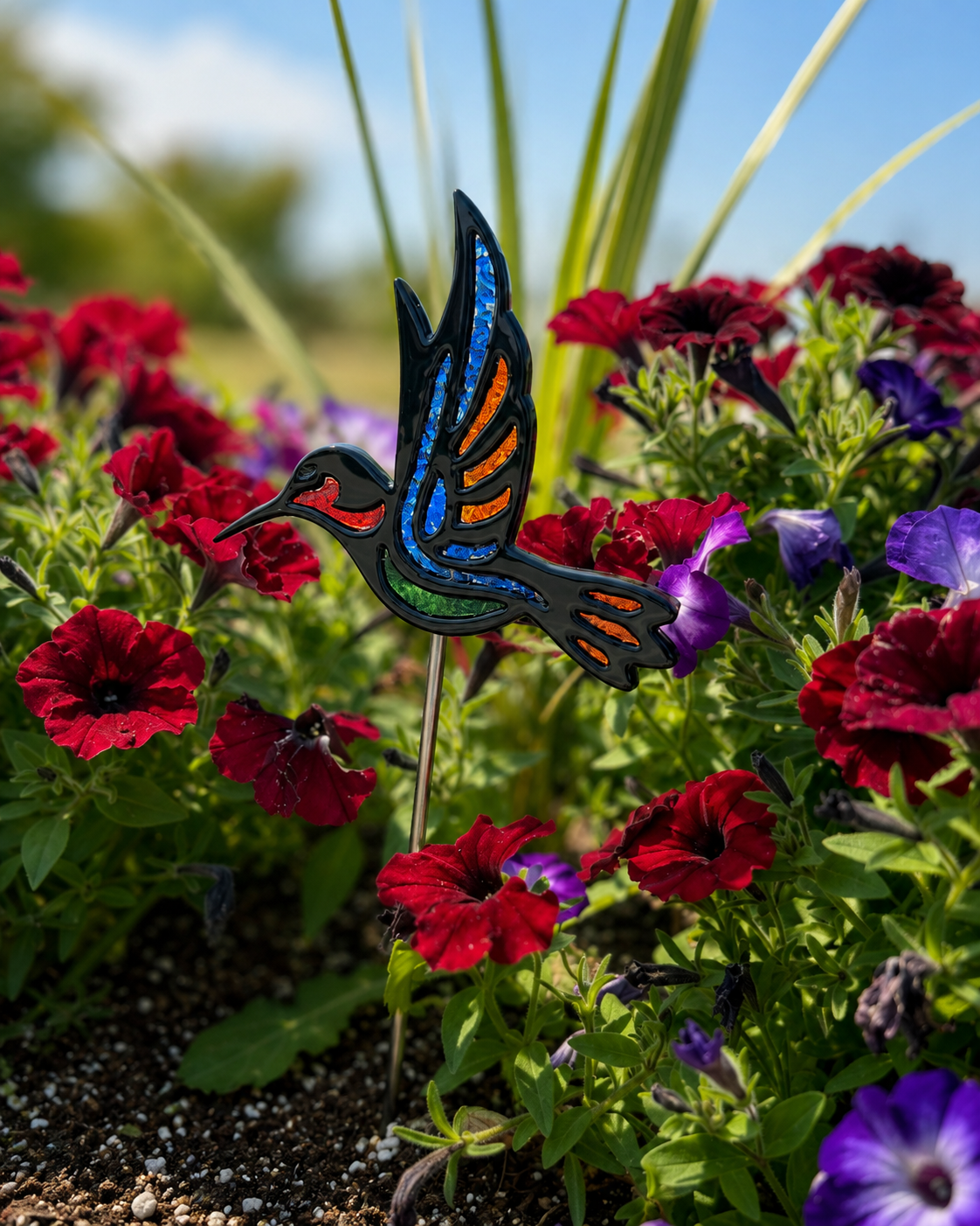 Fused glass hummingbird garden stake with blue, orange and green frit accents displayed among red flowers.