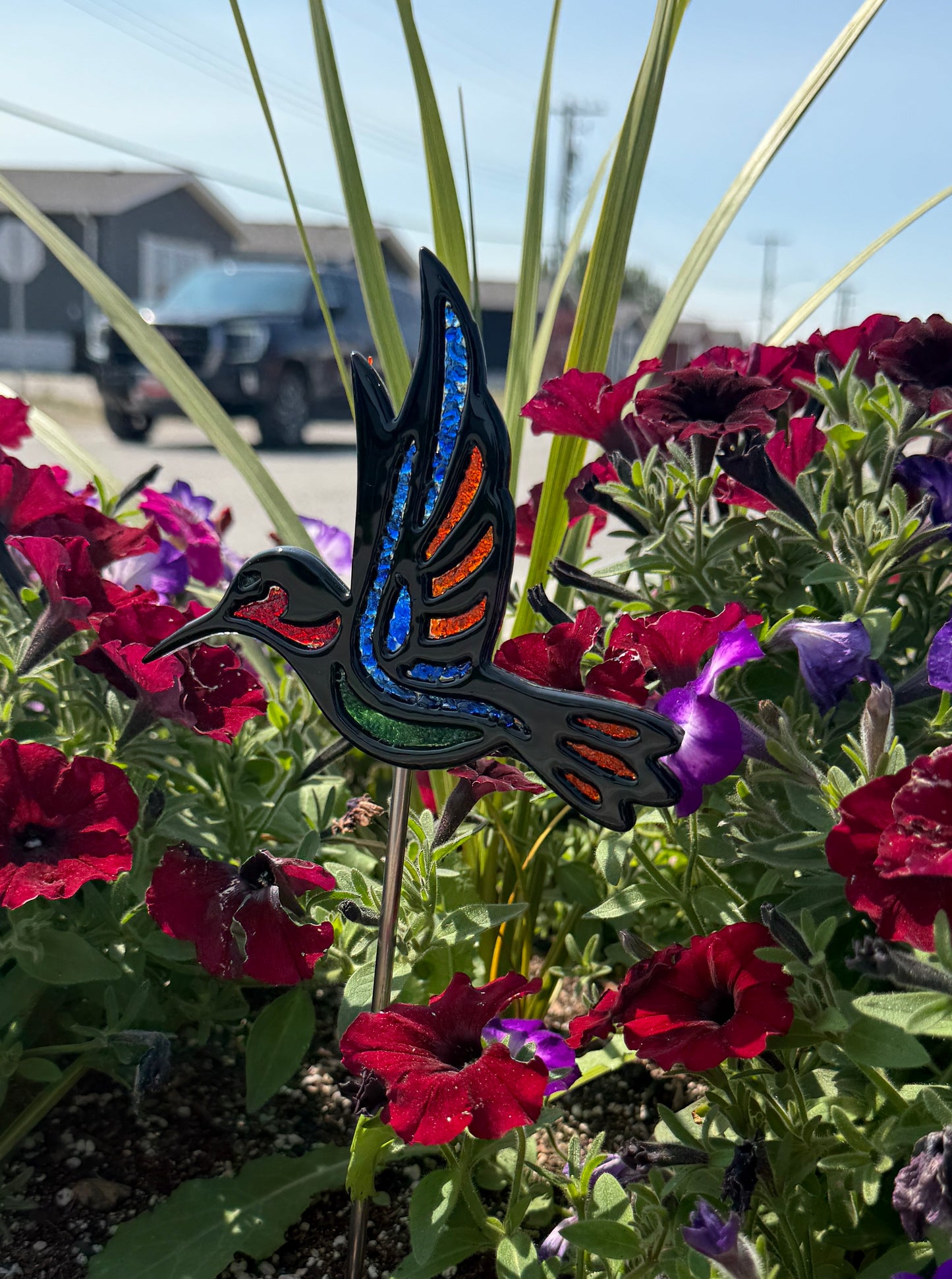 Finished fused glass hummingbird garden stake with blue, green and orange frit accents displayed among red flowers.