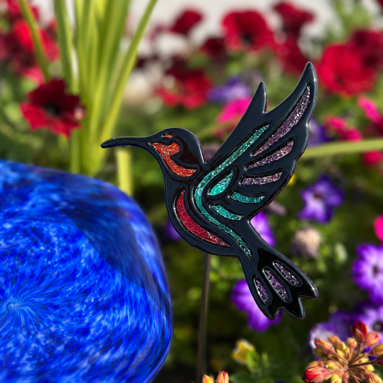 Finished fused glass hummingbird garden stake with black outline and colourful frit accents displayed among flowers.