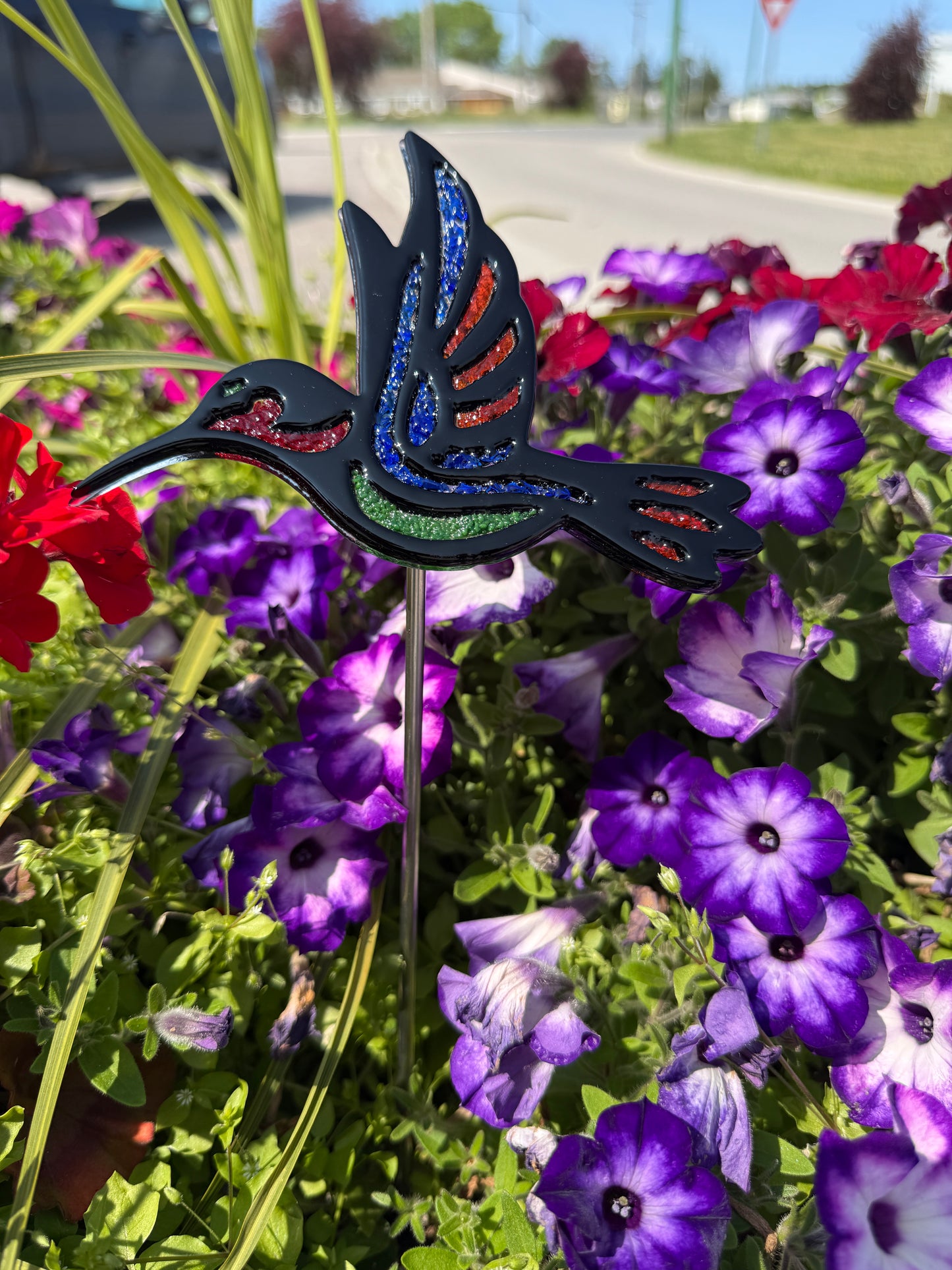 Fused glass hummingbird garden stake with blue, red and green frit accents displayed among purple and red flowers.