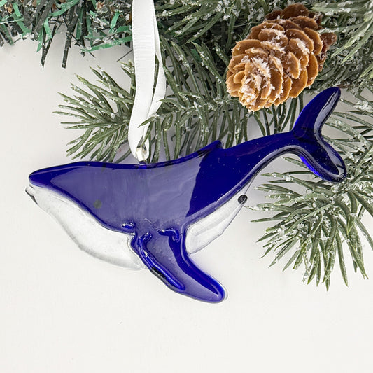 Transparent blue whale Christmas ornament in fused glass, with tree lights sparkling through, ocean-inspired holiday décor.